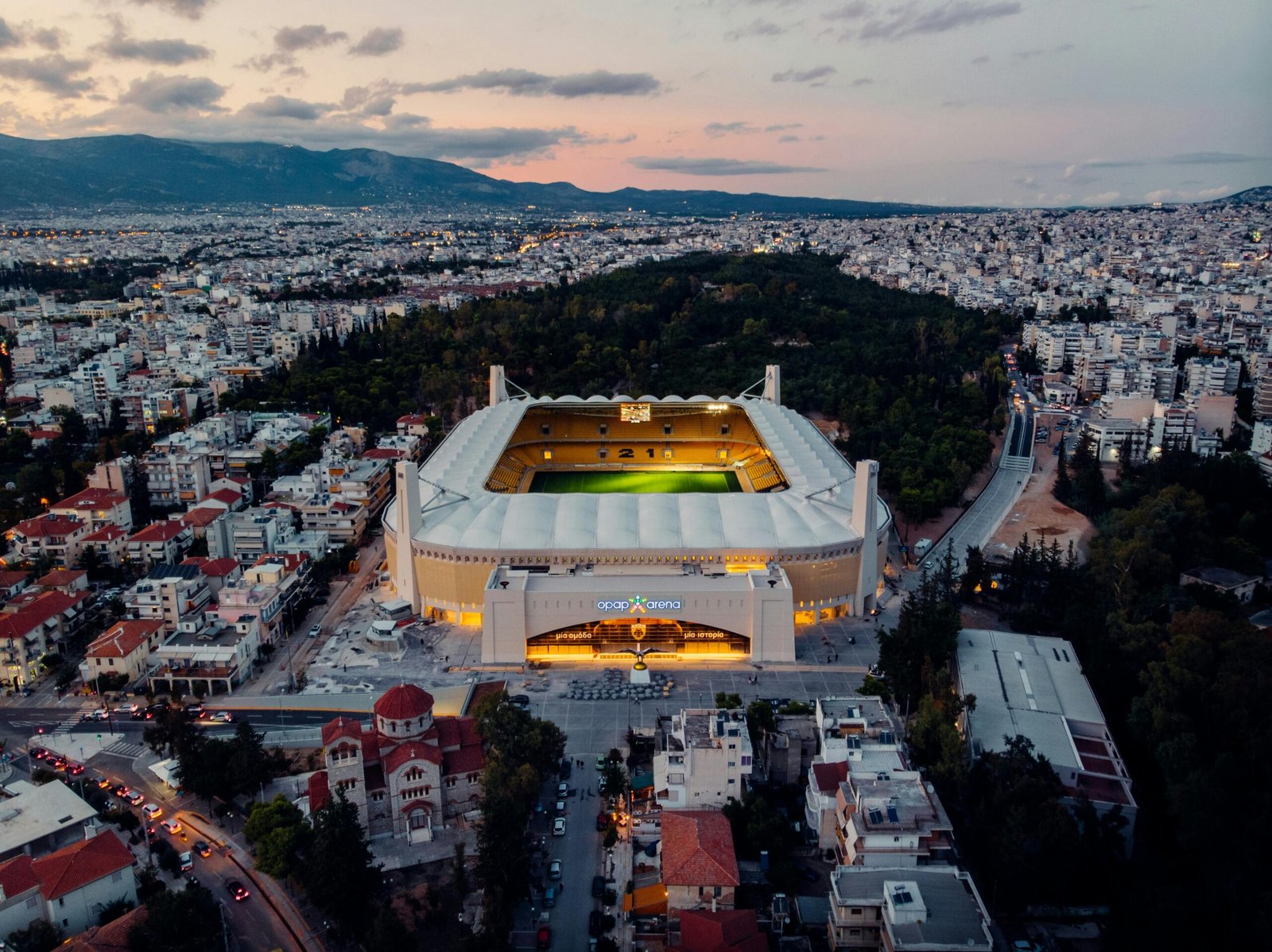 Dramatic aerial shot of OPAP Arena in Athens at twilight, showcasing urban landscape.