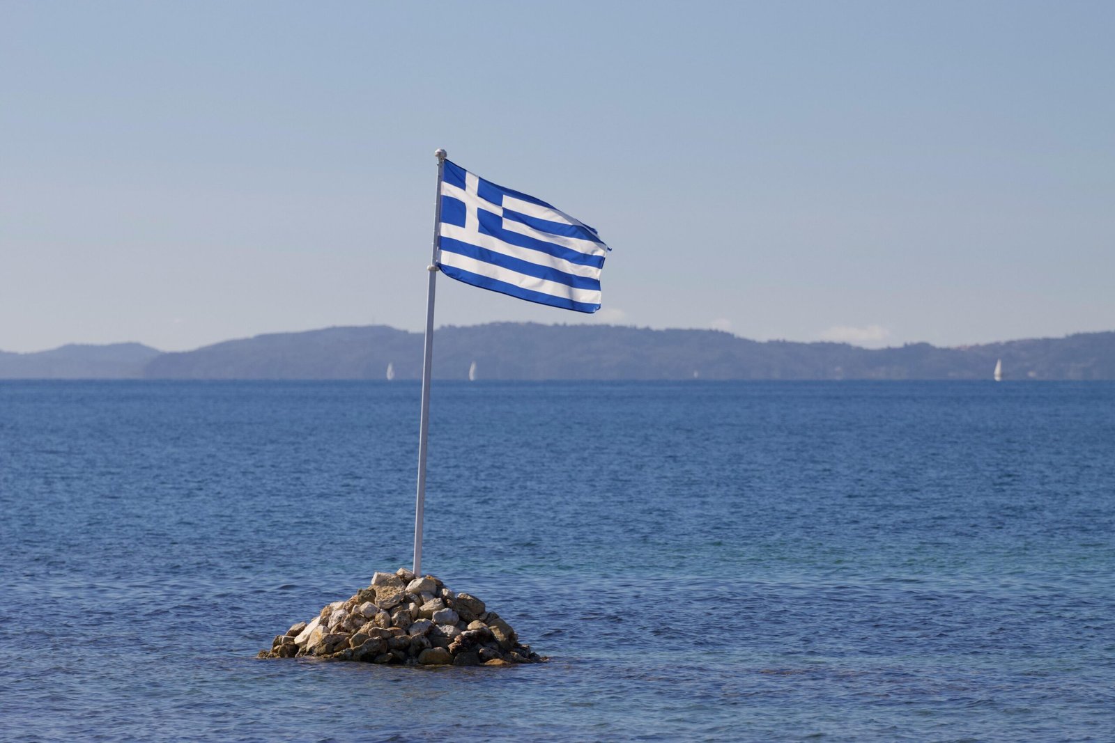 Greek flag waves over rocky island with serene Ionian Sea backdrop in Corfu, Greece.